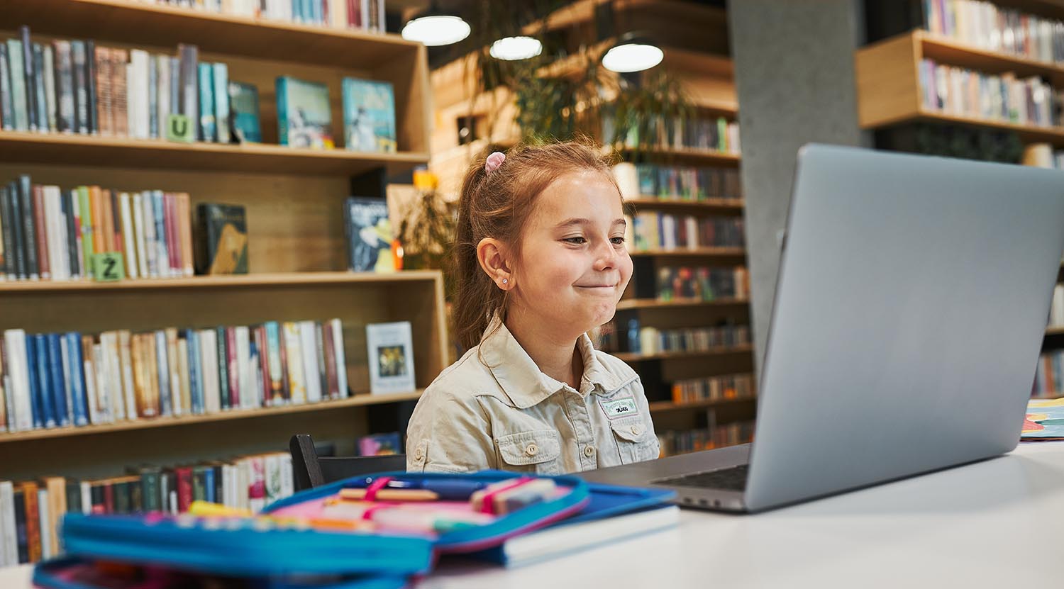 Pleased little student having video class remotely with her teacher on laptop sitting at desk in after school club at primary school. Back to school. Child using technology on elementary computer science lesson. Smart little girl learning online. Teacher